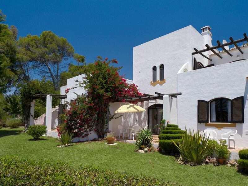 Casa vacanze bianca con pergola circondata da piante verdi e prato ben curato sotto un cielo blu.