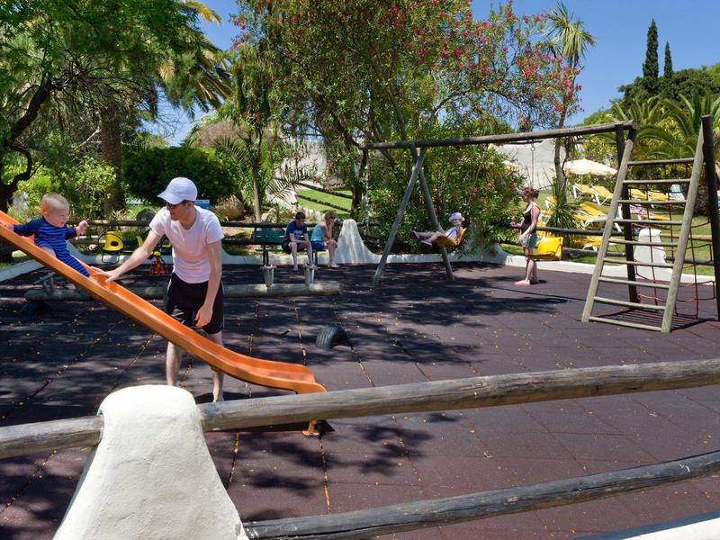 Bambino che gioca su uno scivolo in un parco giochi accanto a un'altalena sotto il sole.