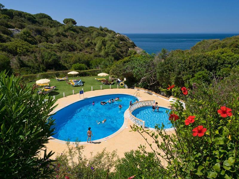 Piscina rotonda con ombrelloni e vista sul mare, circondata da paesaggio verde.
