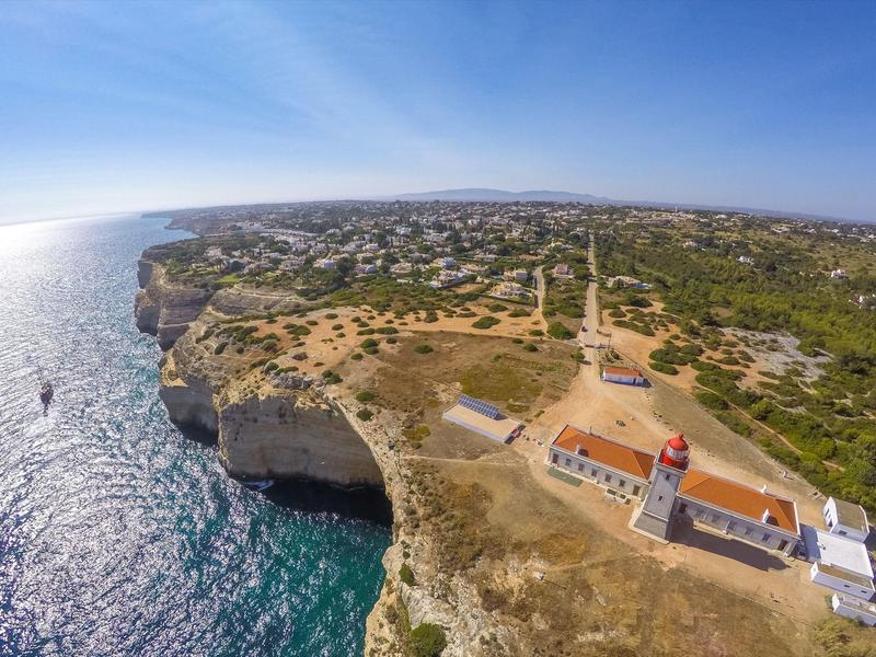 Vista aerea di una scogliera costiera con un faro e il mare sotto un cielo sereno.