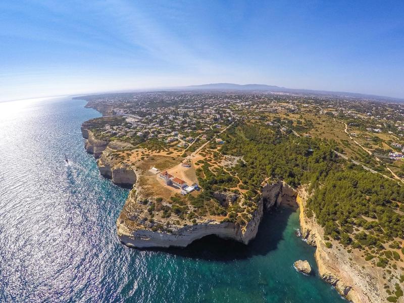 Vista aerea di una costa rocciosa con mare limpido e colline boscose sotto il sole.