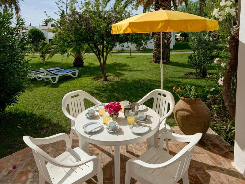 Terrazza con tavolo bianco, sedie e ombrellone giallo in un giardino con lettini.