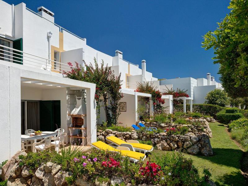 Appartamenti bianchi con terrazze, lettini gialli e giardini verdi sotto un cielo limpido.