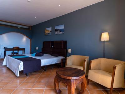 Cozy hotel room with bed, chairs, table, and floor lamp under warm lighting.