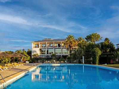 Large outdoor pool in front of a hotel with sun loungers and palm trees under blue sky.