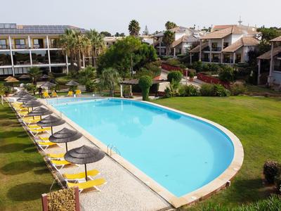 Large pool with umbrellas and loungers in a hotel complex with multiple buildings and garden.