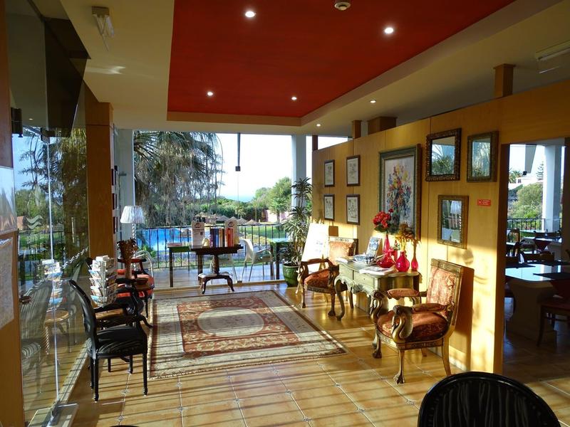 Bright hotel lounge area with armchairs, rugs, and views of the garden and sea.
