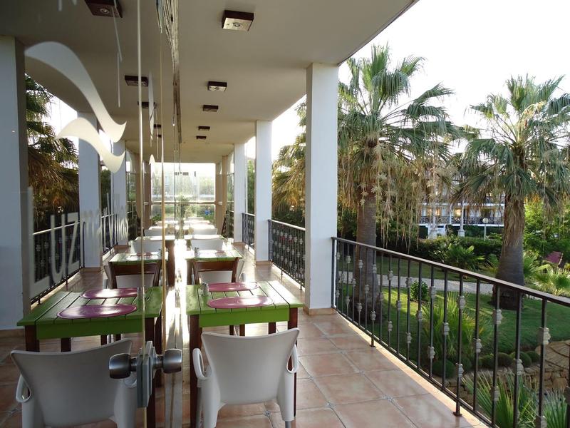 Balcony with tables and chairs next to a garden with palm trees at a hotel.