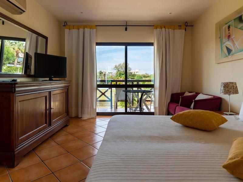 Bright hotel room with double bed, TV on wooden cabinet, and balcony with outdoor view.