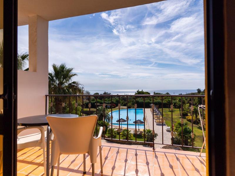 Balcony with chair and table overlooking pool, garden, and sea under blue sky.