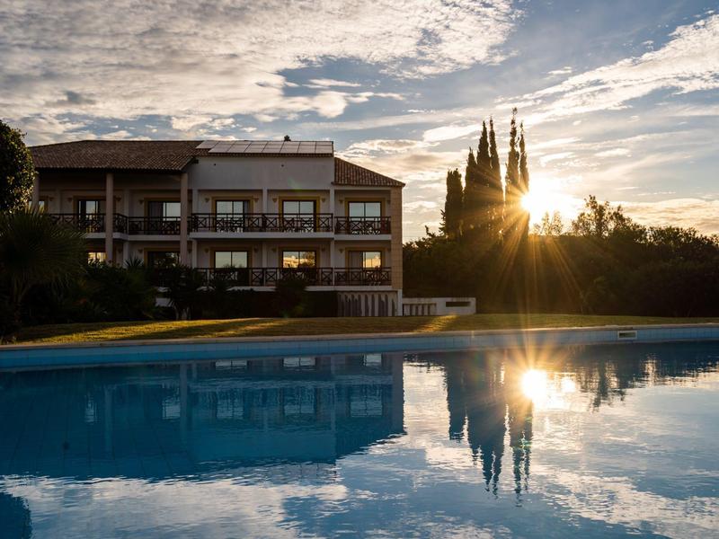 Sunset over a hotel with pool and trees, reflected in the water.