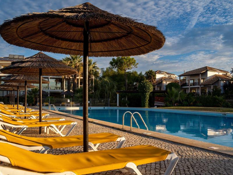 Empty yellow loungers under straw parasols beside a pool under a clear sky.