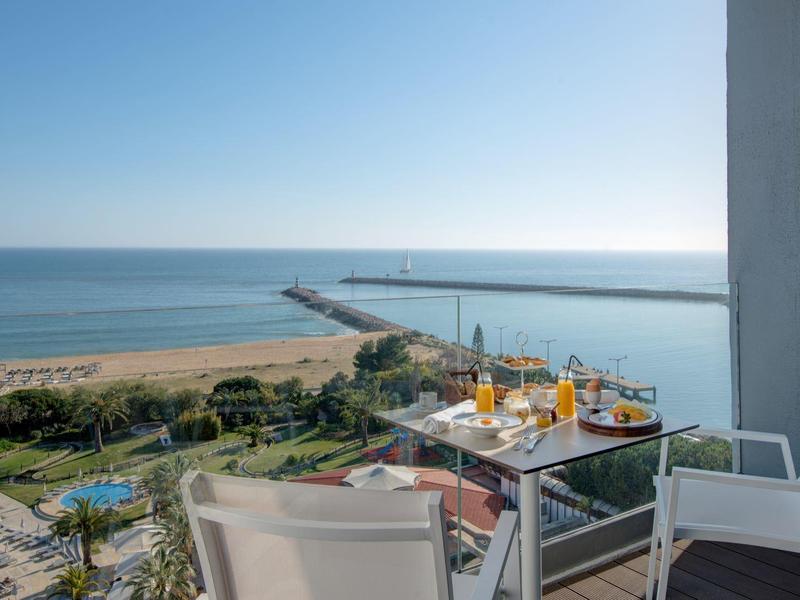 Balkon mit Glas und weißen Stühlen, Tisch mit Getränken, Blick auf Hafen, Strand und Meer bei klarem Himmel.