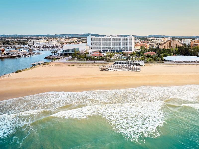 Strand mit goldenem Sand, sanften Wellen und Gebäuden im Hintergrund bei klarem Himmel.
