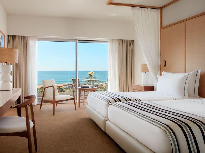 Helles Hotelzimmer mit Doppelbett, modernem Holzmobiliar und großem Fenster mit Meerblick.