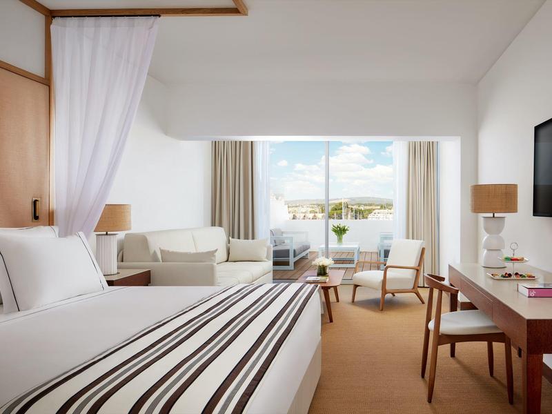 Helles Hotelzimmer mit großem Bett, Schreibtisch, Sessel und Blick auf eine Terrasse mit Wasser.