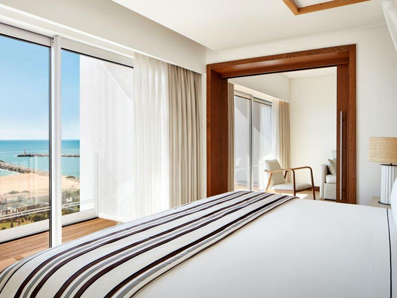 Helles Schlafzimmer mit großem Fenster, Meerblick, Bett mit gestreifter Decke und Holzspiegel.