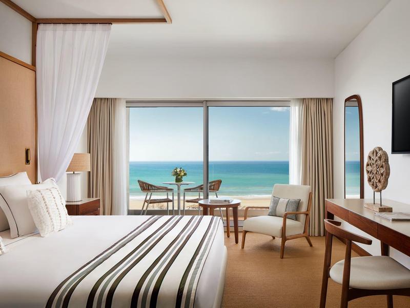 Helles Schlafzimmer mit großem Fenster, Blick aufs Meer, Doppelbett, Tisch, Stuhl und TV an der Wand.