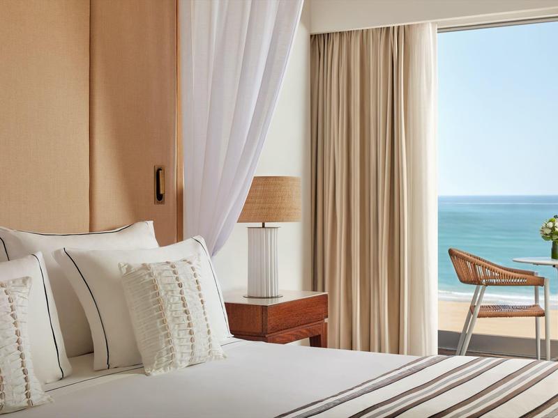 Helles Schlafzimmer mit weißer Bettwäsche, Holzmöbeln und Meerblick durch große Fenster.