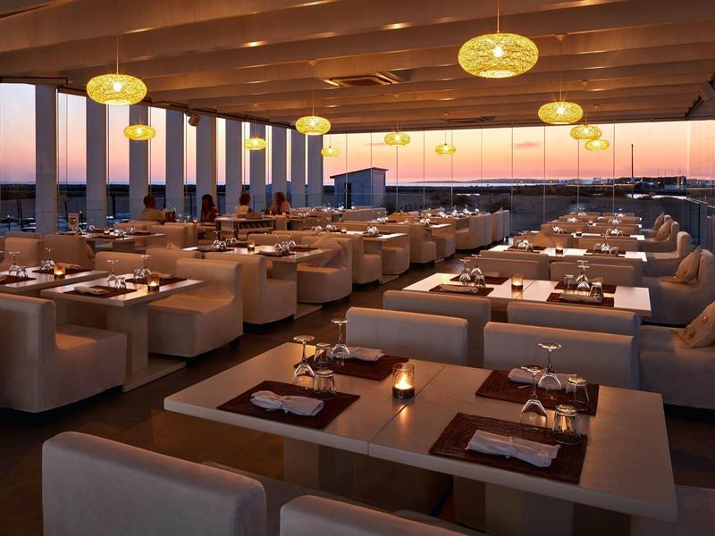 Modernes Restaurant mit braunen Holztischen, beigen Sesseln und gelber Deckenbeleuchtung bei Sonnenuntergang.