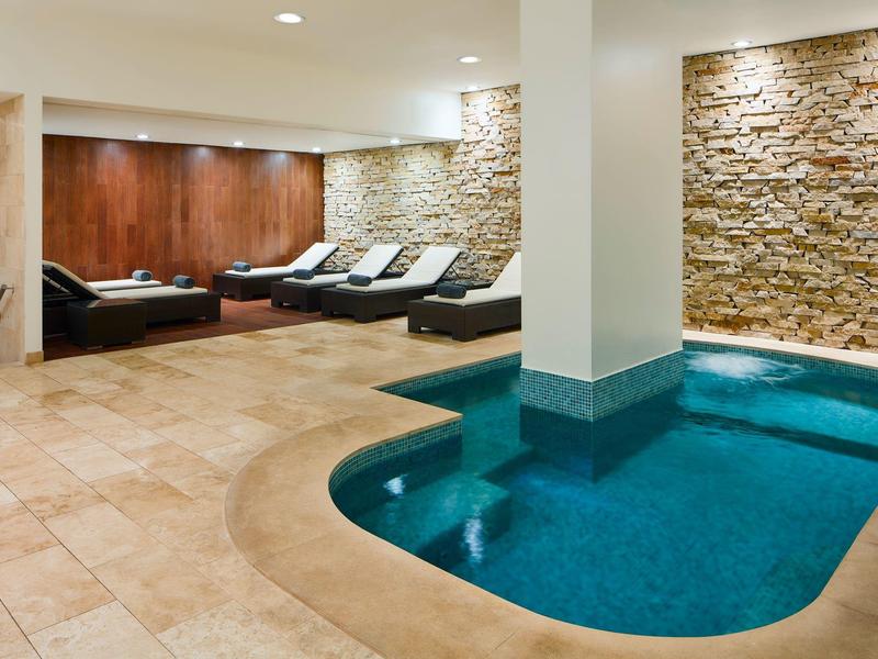 Innenbereich mit kleinem blauen Pool, beige Fliesenboden, Steinsäule und Liegestühlen an Wand.