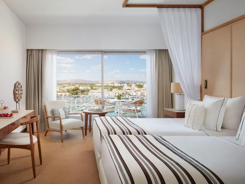 Helles Hotelzimmer mit großem Bett, Schreibtisch, Sesseln und Fenster mit Blick auf Hafen und Stadt.