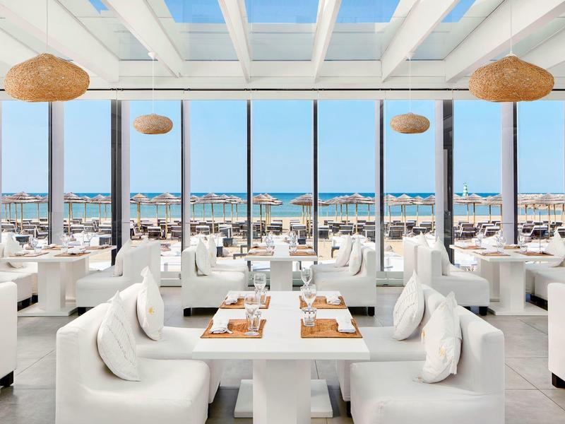 Modernes Restaurant mit weißer Einrichtung und Panoramafenstern mit Blick auf Meer und Strand.