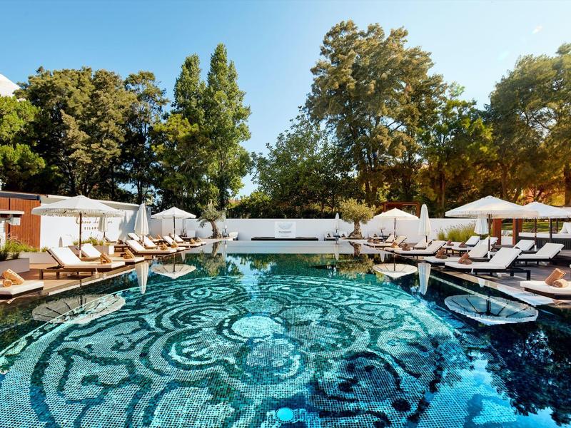 Runder Pool mit Mosaikmuster, umgeben von Liegen und weißen Sonnenschirmen unter Bäumen.