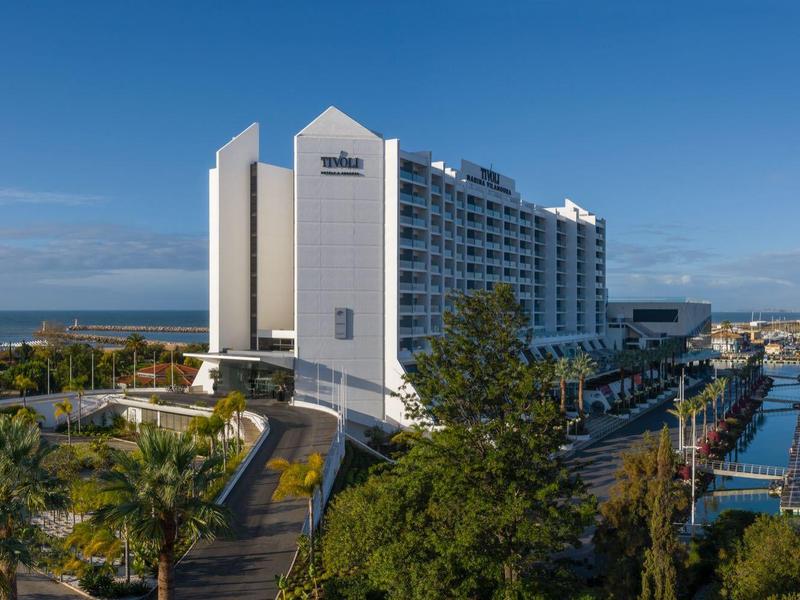 Großes weißes Hotelgebäude am Wasser mit Boots- und Palmenlandschaft unter blauem Himmel.