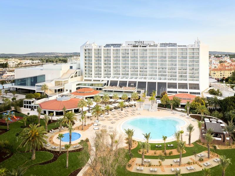 Großes Hotel mit mehreren Pools, umgeben von Palmen und Liegestühlen unter blauem Himmel.