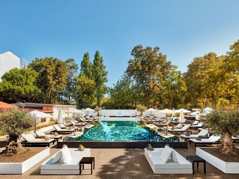 Moderner Poolbereich mit Liegestühlen, weißen Sitzgruppen, Bäumen und klarem blauem Himmel.