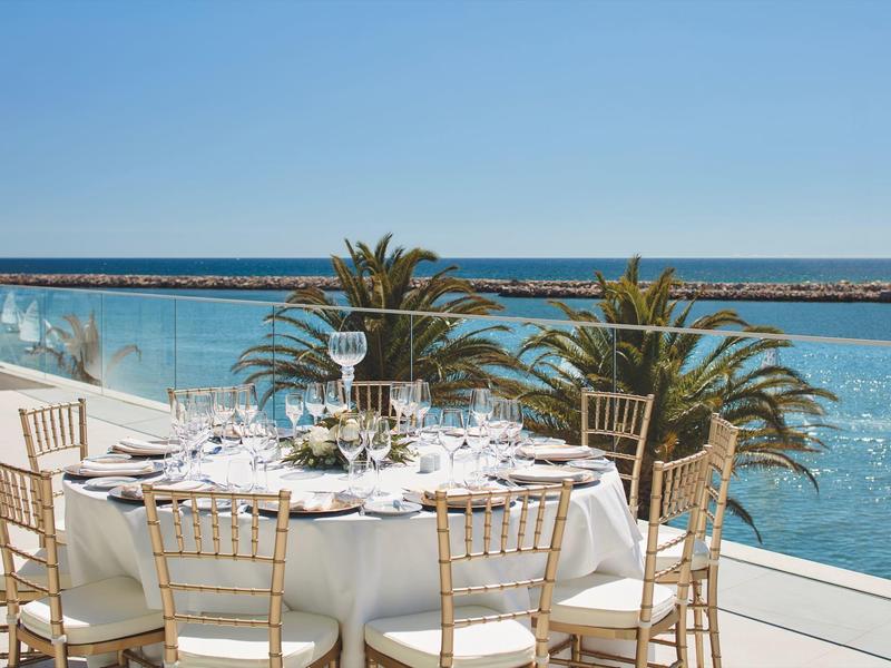 Runder gedeckter Tisch mit Gläsern und Tellern auf Terrasse, Palmen und ruhiges Meer im Hintergrund.