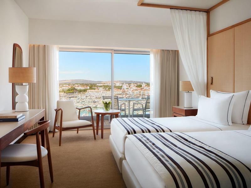 Modernes Hotelzimmer mit zwei Betten, Schreibtisch, Stühlen und großem Fenster mit Stadtblick.