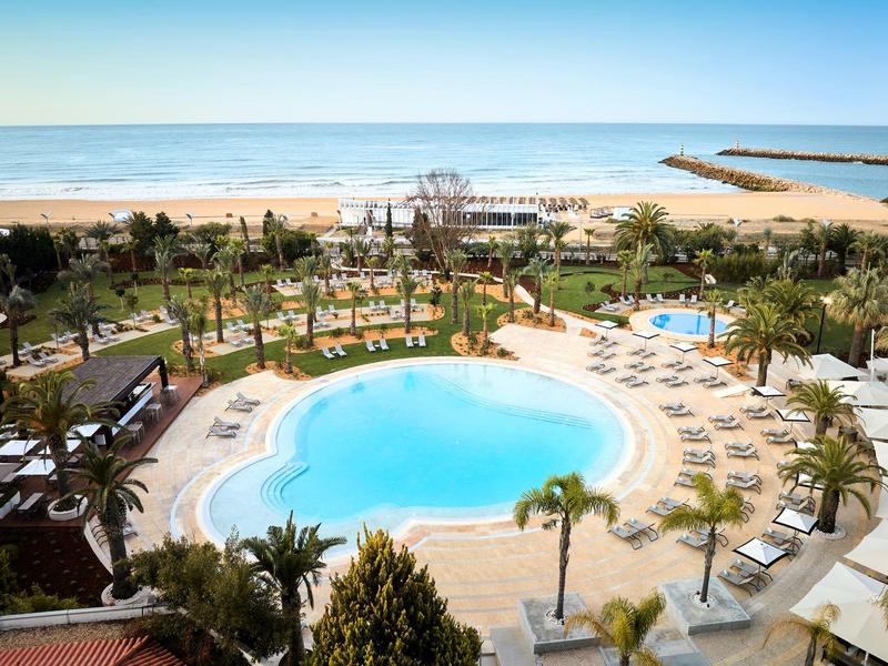 Großer Pool mit Liegestühlen, Palmen und Blick auf Sandstrand und Meer im Hintergrund.