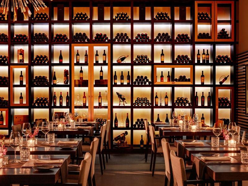 Elegantes Restaurant mit Holztischen, Kerzenlicht und beleuchtetem Weinregal an der Wand.