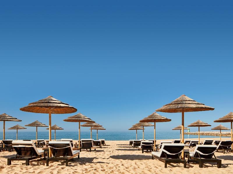 Strand mit Sand, Liegestühlen und Strohmsonnenschirmen unter klarem blauem Himmel.