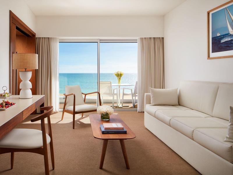 Helles Wohnzimmer mit Sofa, Holzstuhl, Tisch, Meerblick und Balkon mit Glasgeländer.