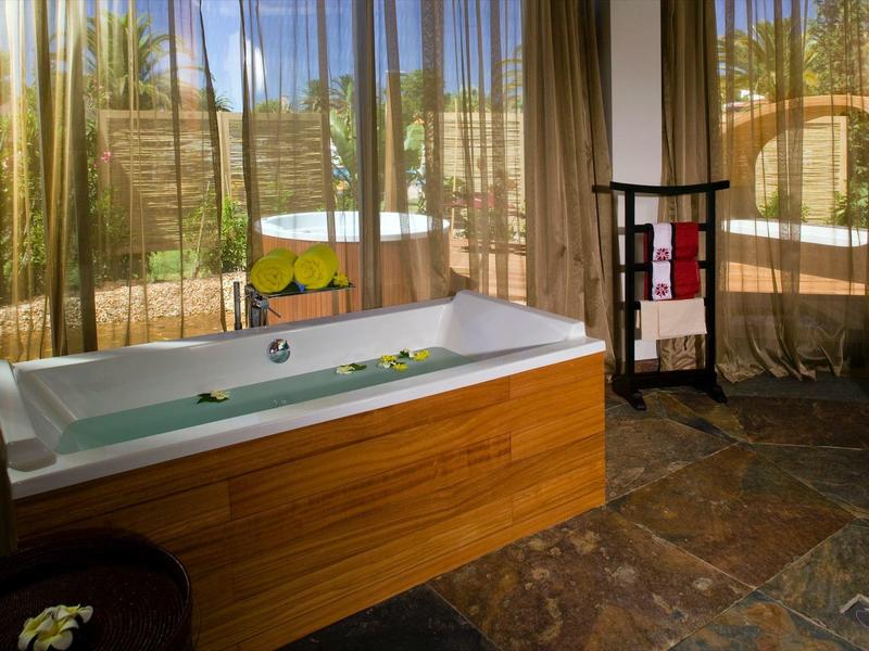 Helles Bad mit freistehender Holzbadewanne, Fenster mit Vorhängen und Steinboden im warmen Licht.