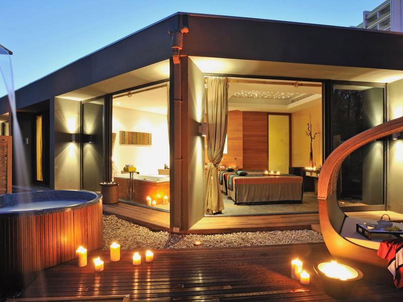 Moderner Außenbereich mit Holzterrasse, Hot Tub, brennenden Kerzen und offenem, beleuchtetem Wohnraum.