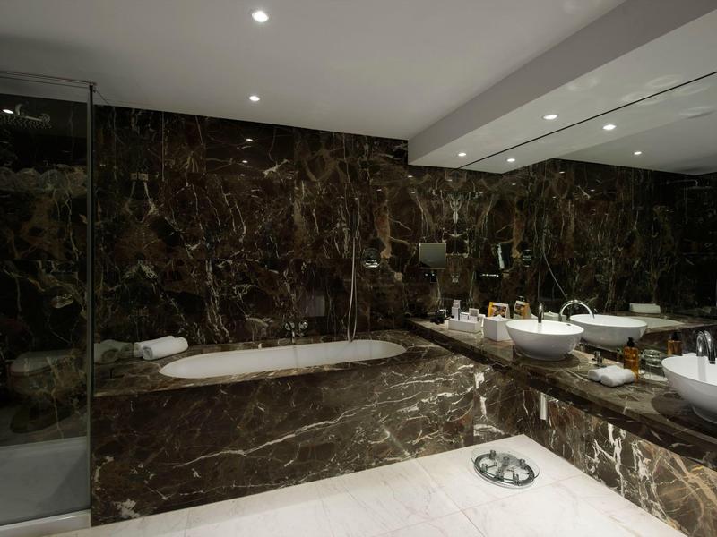 Luxuriöses Badezimmer mit dunkelbraunen Marmorwänden, weißer Badewanne und Waschbecken, Spiegeln und Beleuchtung.