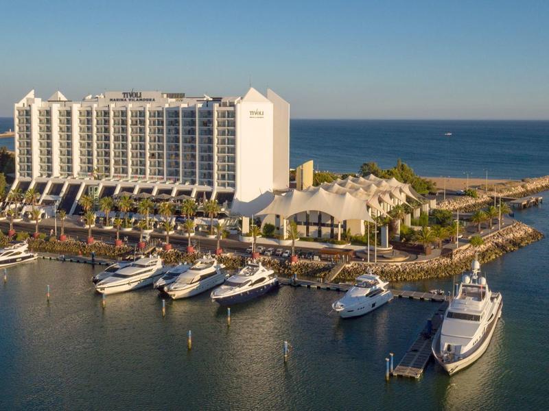 Großes Hotelgebäude an der Küste mit mehreren Yachten im Hafen und blauem Meer im Hintergrund.
