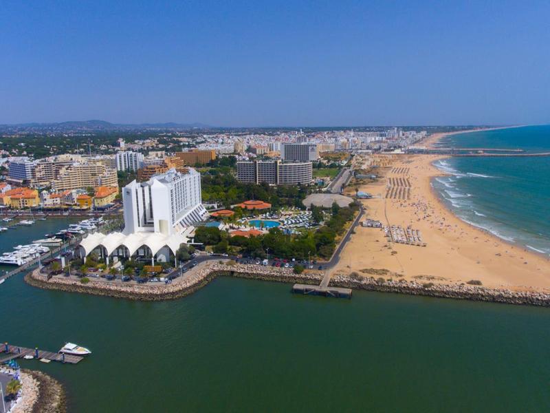 Luftaufnahme einer Küstenstadt mit breitem Sandstrand, Hafen und zahlreichen Gebäuden bei klarem Himmel.