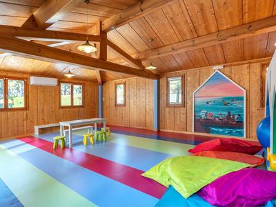 Stanza giochi luminosa con pavimento colorato, tavoli piccoli e cuscini sotto travi di legno.