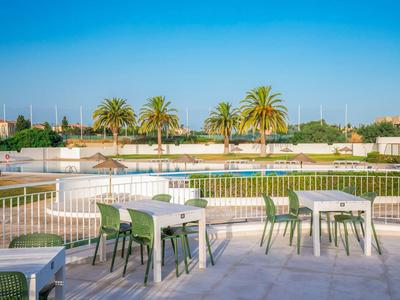 Terrasse mit weißen Tischen und grünen Stühlen, Palmen und klarer blauer Himmel im Hintergrund.