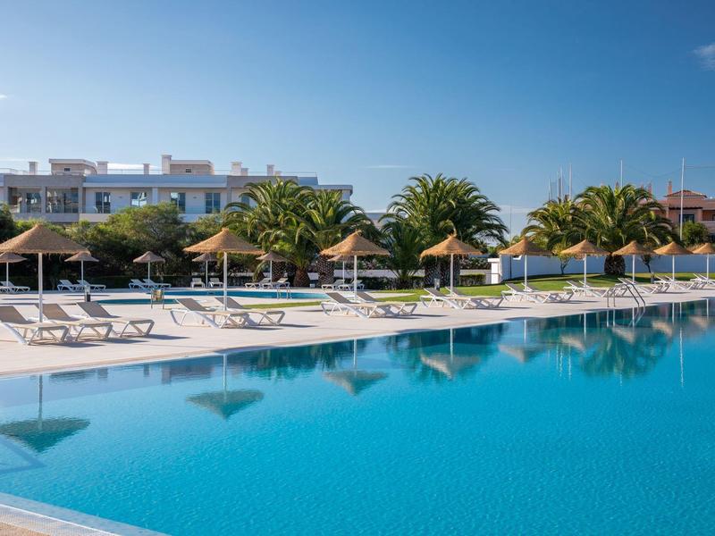 Duży basen z leżakami i parasolami przed budynkami hotelu i palmami pod bezchmurnym niebem.