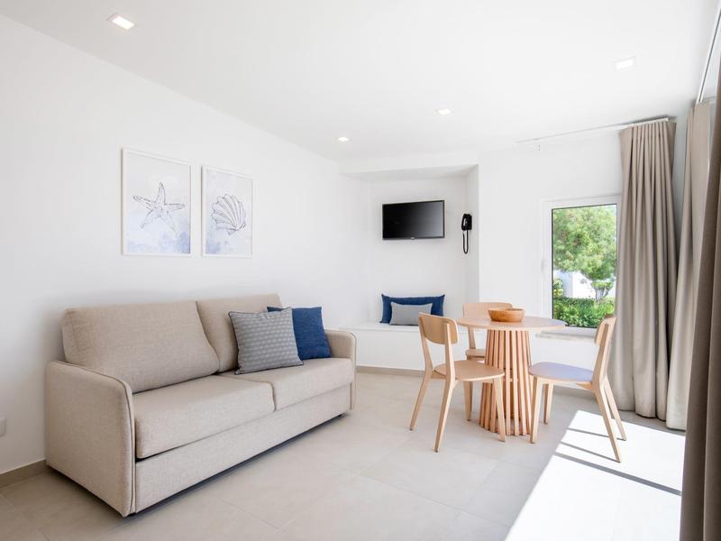 Jasny, nowoczesny pokój hotelowy z sofą, stołem jadalnym, krzesłami i telewizorem na ścianie.