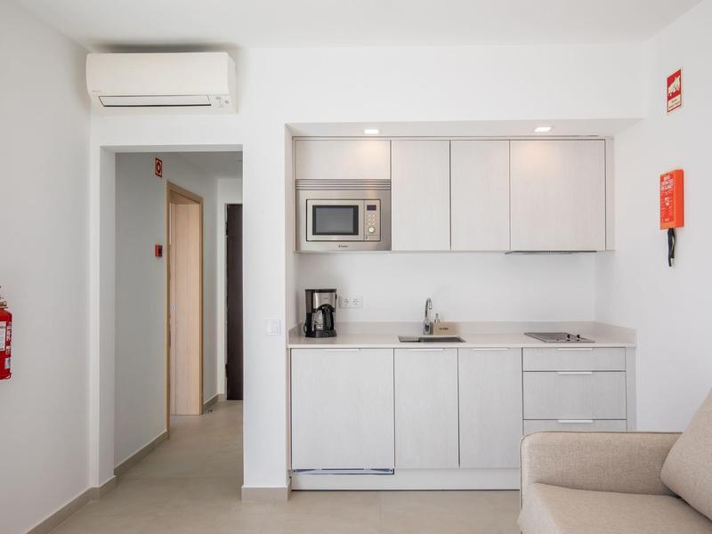 Kompaktowa biała kuchnia z klimatyzacją i beżową sofą w jasnym pomieszczeniu.