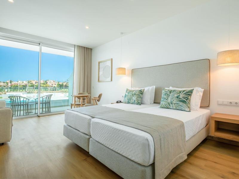 Jasny pokój hotelowy z dużym oknem, widokiem na wodę i balkonem z krzesłami.