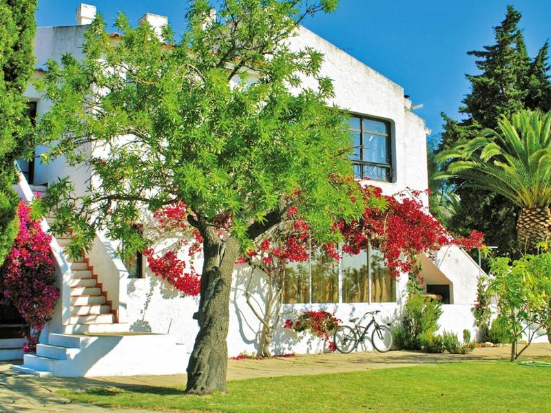 Casa de vacaciones blanca con flores rojas, césped verde y cielo azul al fondo.