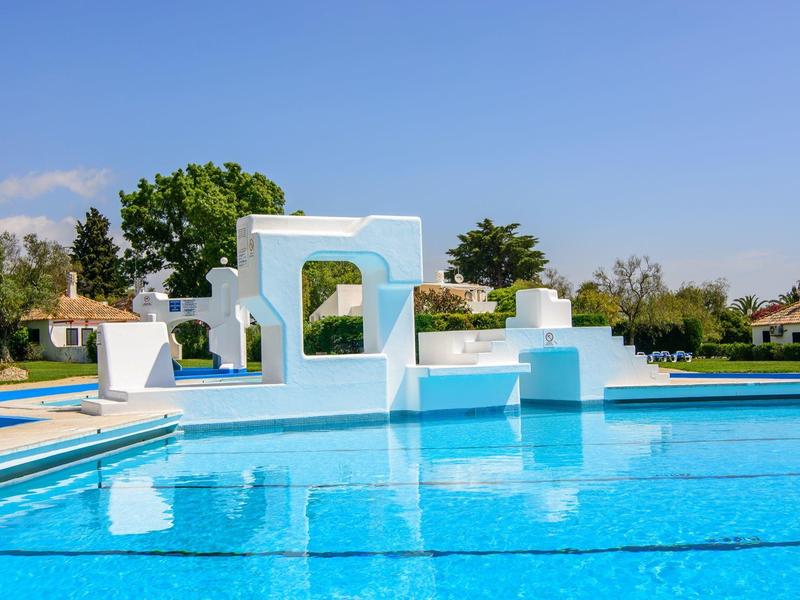 Gran piscina azul con trampolines blancos y zonas para tomar el sol bajo un cielo despejado.